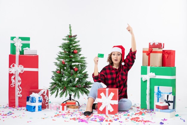 Vue de face de la jeune femme assise autour de cadeaux tenant une carte bancaire verte sur un mur blanc