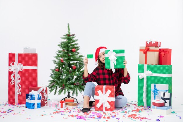 Vue de face de la jeune femme assise autour de cadeaux tenant une carte bancaire verte et présente sur mur blanc