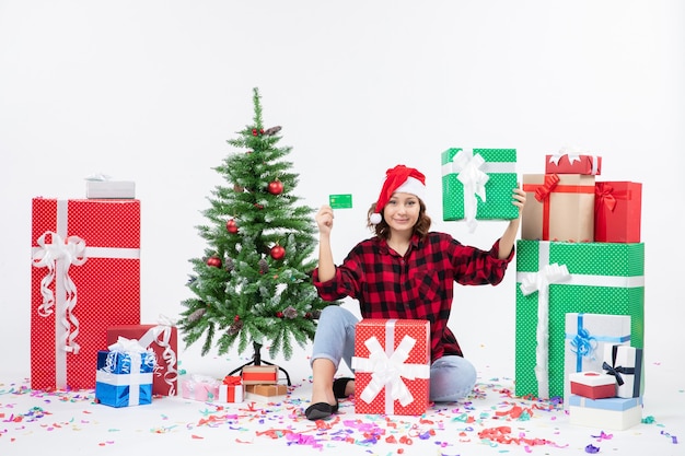 Vue de face de la jeune femme assise autour de cadeaux tenant une carte bancaire verte et présente sur mur blanc