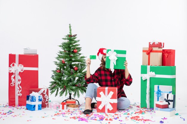 Vue de face de la jeune femme assise autour de cadeaux tenant une carte bancaire verte et présente sur mur blanc