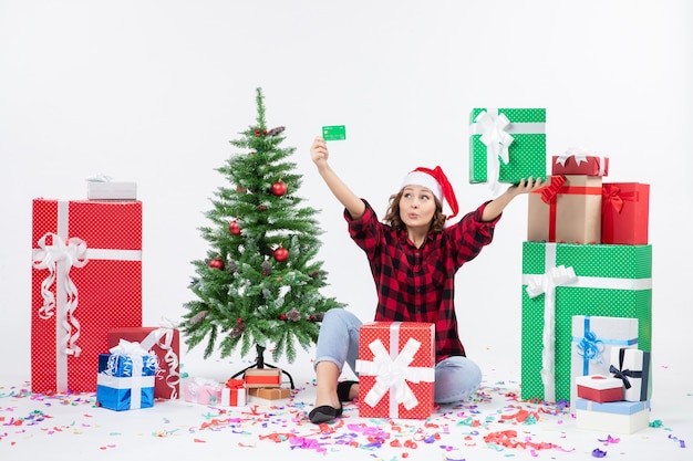 Vue de face de la jeune femme assise autour de cadeaux tenant une carte bancaire verte et présente sur mur blanc