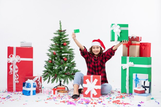 Vue de face de la jeune femme assise autour de cadeaux tenant une carte bancaire verte et présente sur mur blanc