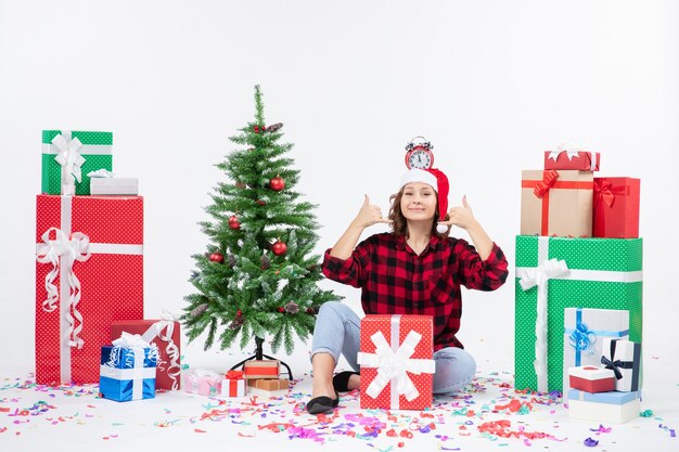 Vue de face de la jeune femme assise autour de cadeaux tenant des horloges sur sa tête sur un mur blanc