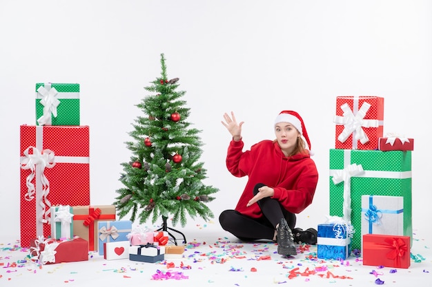 Vue de face de la jeune femme assise autour des cadeaux de vacances sur le mur blanc