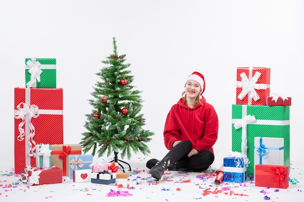 Vue de face de la jeune femme assise autour des cadeaux de vacances sur le mur blanc