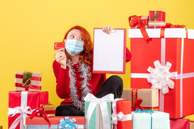 Vue de face de la jeune femme assise autour de Noël présente en masque avec carte bancaire sur mur jaune