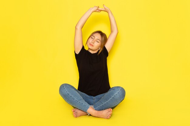 Une vue de face jeune femme assise en chemise noire et blue-jeans faisant de la gymnastique sur jaune