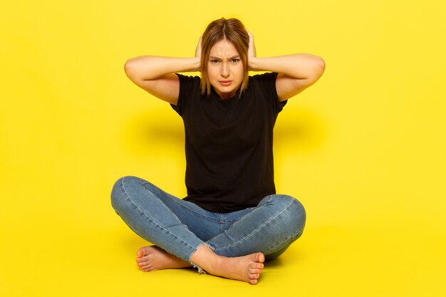 Une vue de face jeune femme assise en chemise noire et un jean bleu couvrant ses oreilles sur jaune