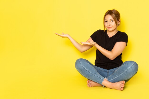 Une vue de face jeune femme assise en chemise noire et jean bleu posant et soulignant avec sourire sur jaune