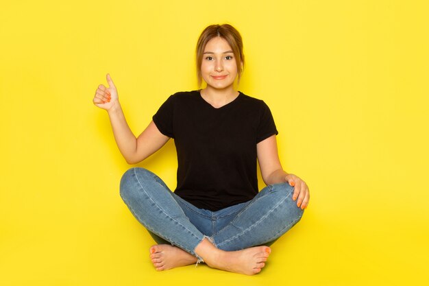 Une vue de face jeune femme assise en chemise noire et jean bleu posant avec sourire sur jaune