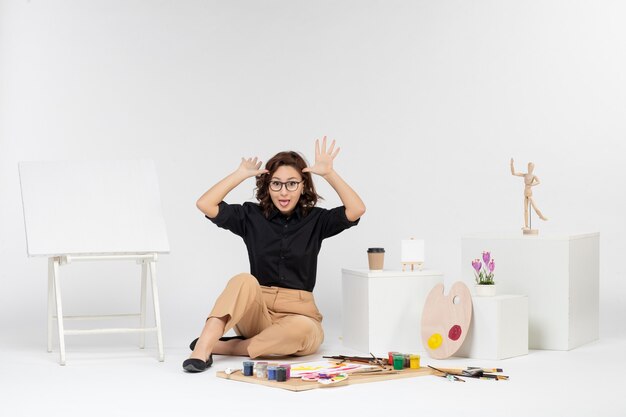 Vue de face jeune femme assise à l'intérieur de la chambre avec chevalet et peintures sur l'art du bureau blanc dessiner image peintre dessin artiste couleur