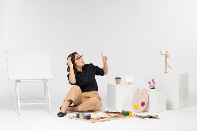 Vue de face jeune femme assise à l'intérieur de la chambre avec chevalet et peintures sur fond blanc