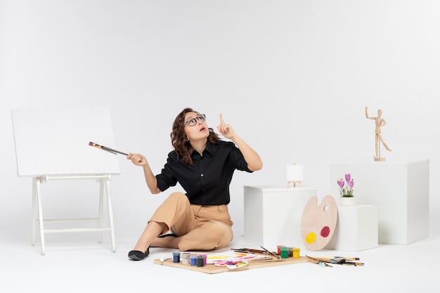 Photo gratuite vue de face jeune femme assise à l'intérieur de la pièce avec des peintures et un chevalet sur fond blanc