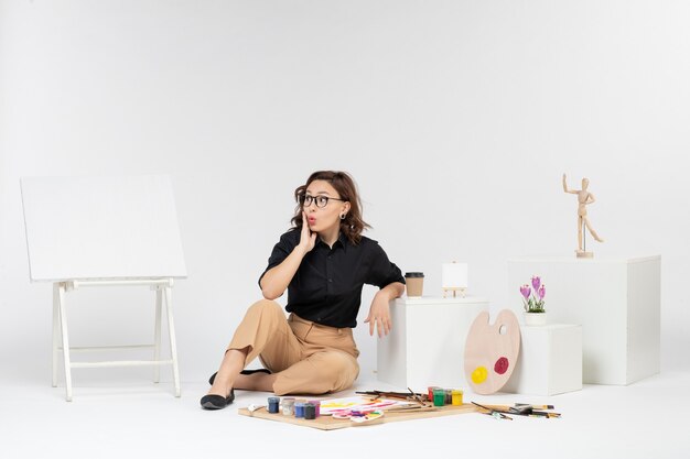 Vue de face jeune femme assise à l'intérieur de la salle avec des peintures pompon et chevalet sur fond blanc