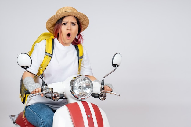 Vue de face jeune femme assise sur une moto avec sac à dos et chapeau mur blanc