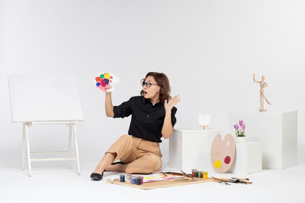 Vue de face jeune femme assise avec des peintures et un chevalet sur un bureau blanc couleur peintre dessin artiste art femme dessiner