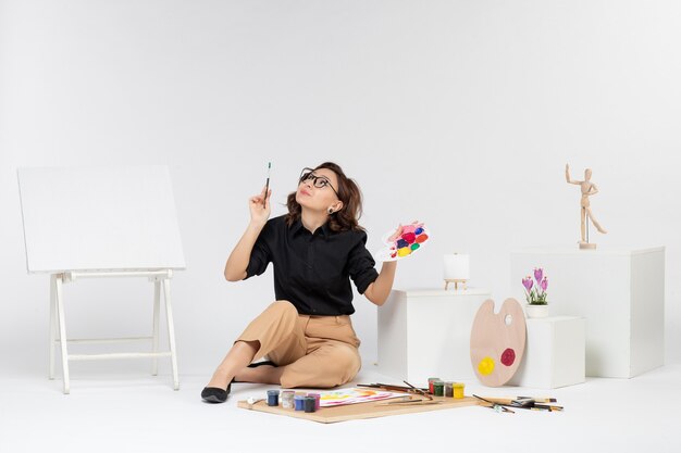 Vue de face jeune femme assise avec des peintures et un chevalet sur un bureau blanc peintre couleur dessin artiste art femme dessiner