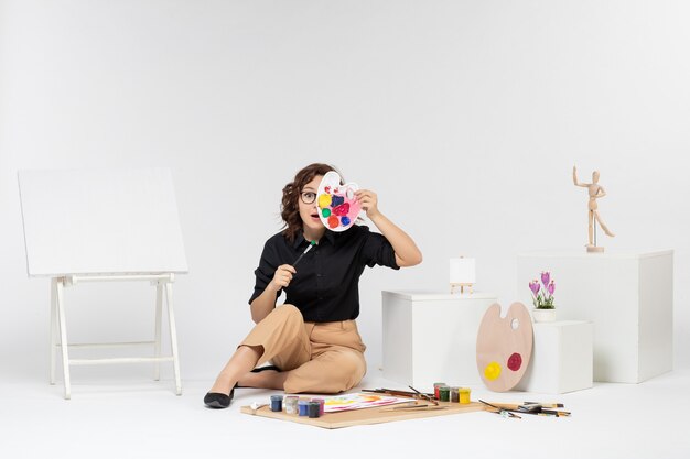 Vue de face jeune femme assise avec des peintures et chevalet sur fond blanc