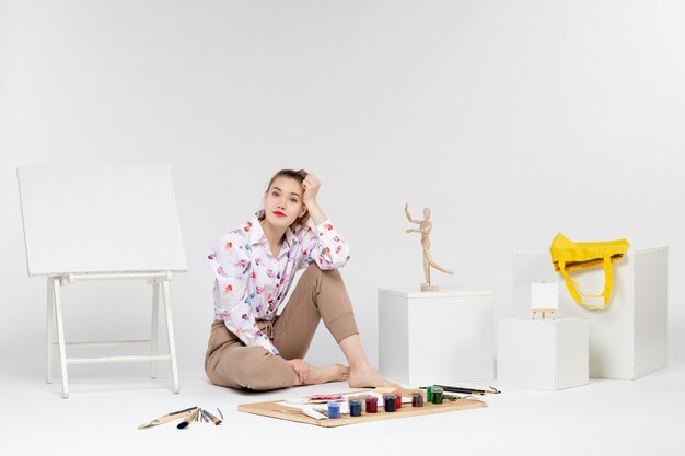 Vue de face jeune femme assise avec des peintures chevalet et pinceaux sur fond blanc