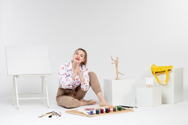 Vue de face jeune femme assise avec des peintures chevalet et pinceaux sur fond blanc