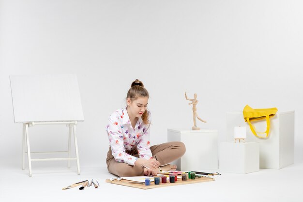 Vue de face jeune femme assise avec des peintures et un chevalet pour dessiner sur un bureau blanc art dessiner femme peintre chevalet artiste couleur