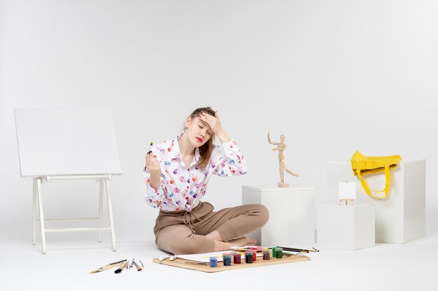 Vue de face jeune femme assise avec des peintures et un chevalet pour dessiner sur un chevalet de bureau blanc artiste femme peintre couleur art dessiner