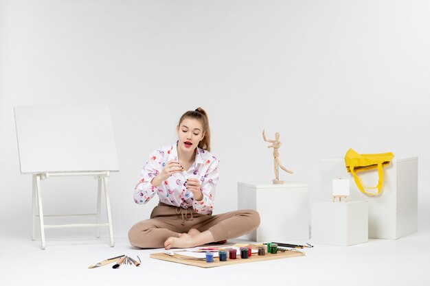 Vue de face jeune femme assise avec des peintures et un chevalet pour dessiner sur fond blanc