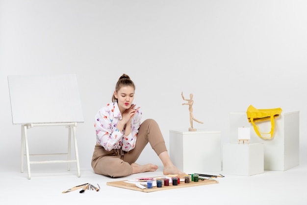 Vue de face jeune femme assise avec des peintures et un chevalet pour dessiner sur fond blanc