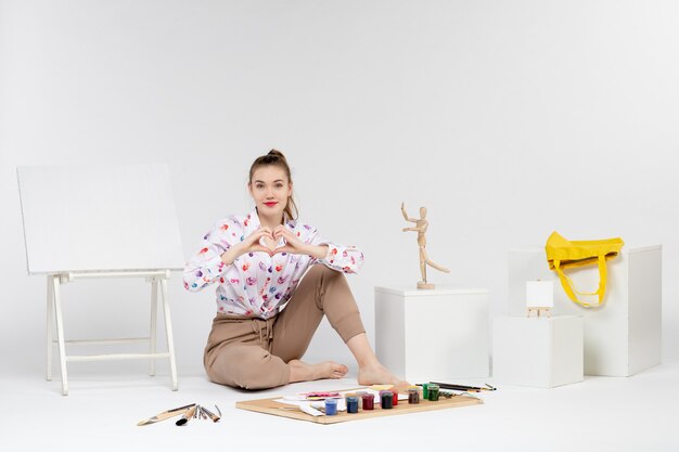 Vue de face jeune femme assise avec des peintures et un chevalet pour dessiner sur fond blanc