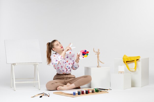 Vue de face jeune femme assise avec des peintures et un chevalet pour dessiner sur fond blanc