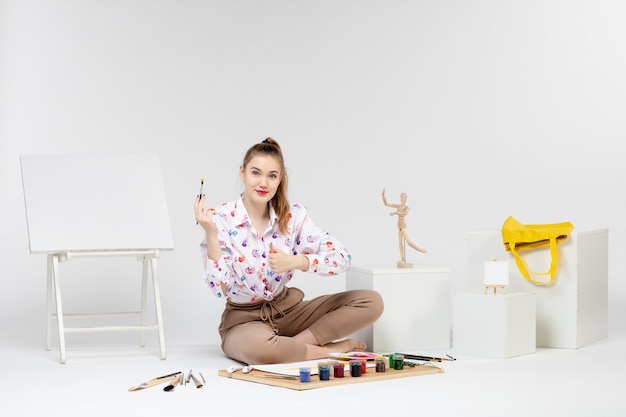 Vue de face jeune femme assise avec des peintures et un chevalet pour dessiner sur fond blanc