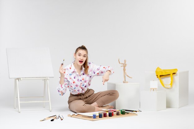 Vue de face jeune femme assise avec des peintures et un chevalet pour dessiner sur fond blanc