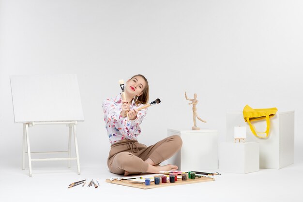 Vue de face jeune femme assise avec des peintures et un chevalet pour dessiner sur fond blanc