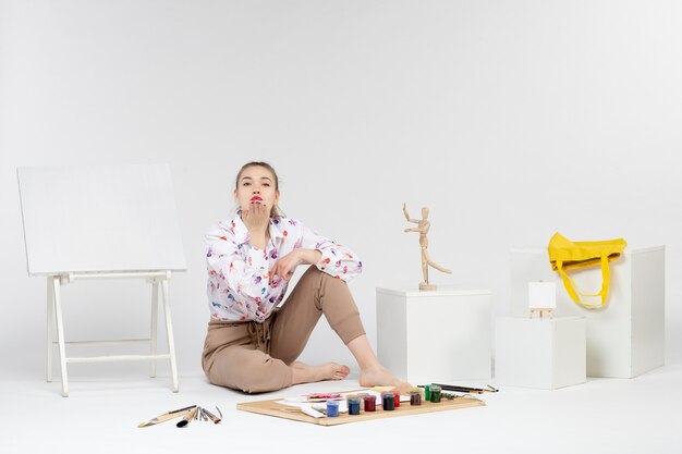 Vue de face jeune femme assise avec des peintures et un chevalet pour dessiner sur fond blanc