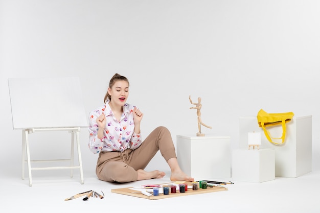 Vue de face jeune femme assise avec des peintures et un chevalet pour dessiner sur fond blanc