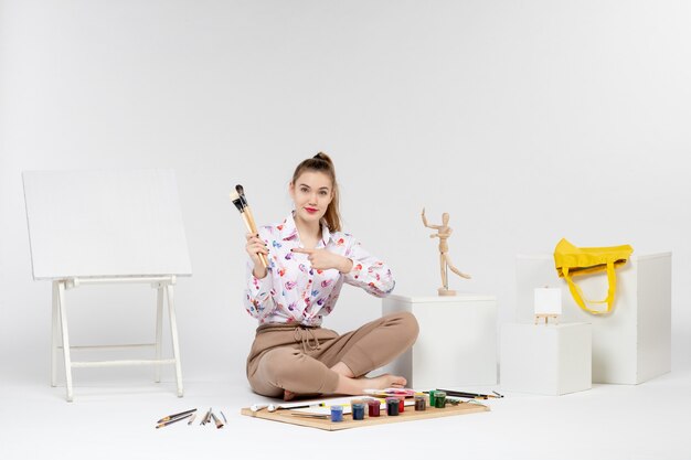 Vue de face jeune femme assise avec des peintures tenant des pinceaux sur fond blanc