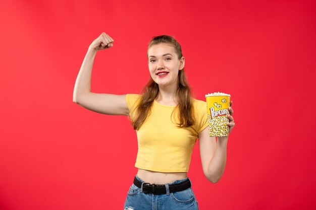 Vue de face jeune femme au cinéma tenant le paquet de pop-corn et fléchissant sur le mur rouge films théâtre cinéma snack féminin film amusant