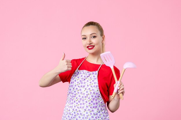 Vue de face jeune femme au foyer avec des couverts dans ses mains sur un mur rose