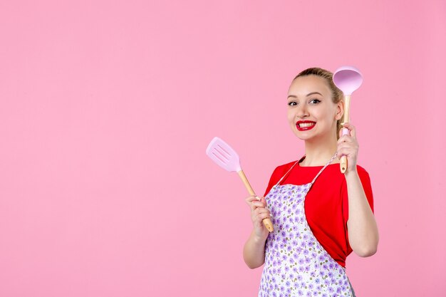 Vue de face jeune femme au foyer posant avec des couverts dans ses mains sur un mur rose