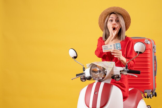 Vue de face jeune femme aux yeux écarquillés en robe rouge tenant un billet sur un cyclomoteur