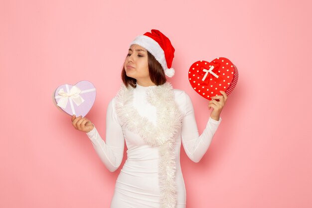 Vue de face d'une jeune femme en bonnet rouge tenant des cadeaux en forme de coeur sur un mur rose