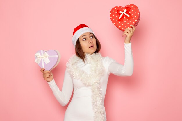 Vue de face d'une jeune femme en bonnet rouge tenant des cadeaux en forme de coeur sur un mur rose