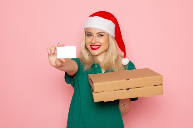 Vue de face d'une jeune femme en bonnet rouge tenant une carte bancaire et des boîtes à pizza sur un mur rose