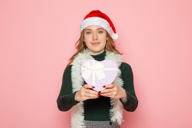 Vue de face d'une jeune femme en bonnet rouge tenant un présent en forme de coeur sur un mur rose