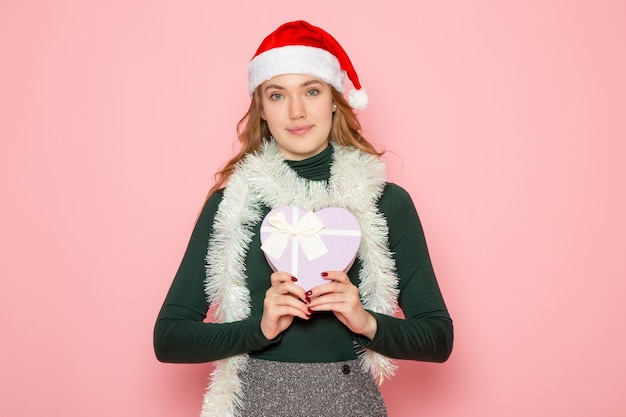 Vue de face d'une jeune femme en bonnet rouge tenant un présent en forme de coeur sur un mur rose