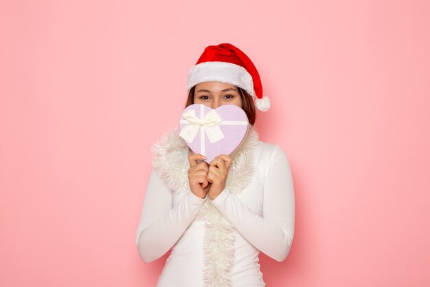 Vue de face d'une jeune femme en bonnet rouge tenant un présent en forme de coeur sur un mur rose