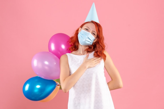 Vue de face jeune femme cachant des ballons colorés derrière son dos dans un masque stérile sur le fond rose partie covid- couleur du nouvel an de Noël