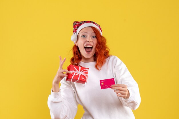 Vue de face de la jeune femme avec cadeau de Noël et carte bancaire sur mur jaune