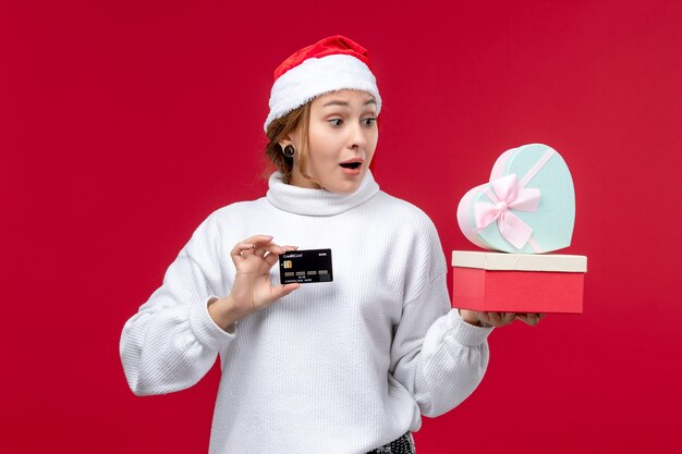 Vue de face jeune femme avec des cadeaux et une carte bancaire sur le fond rouge