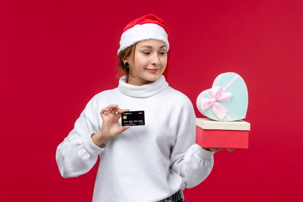 Vue de face jeune femme avec des cadeaux et une carte bancaire sur le fond rouge